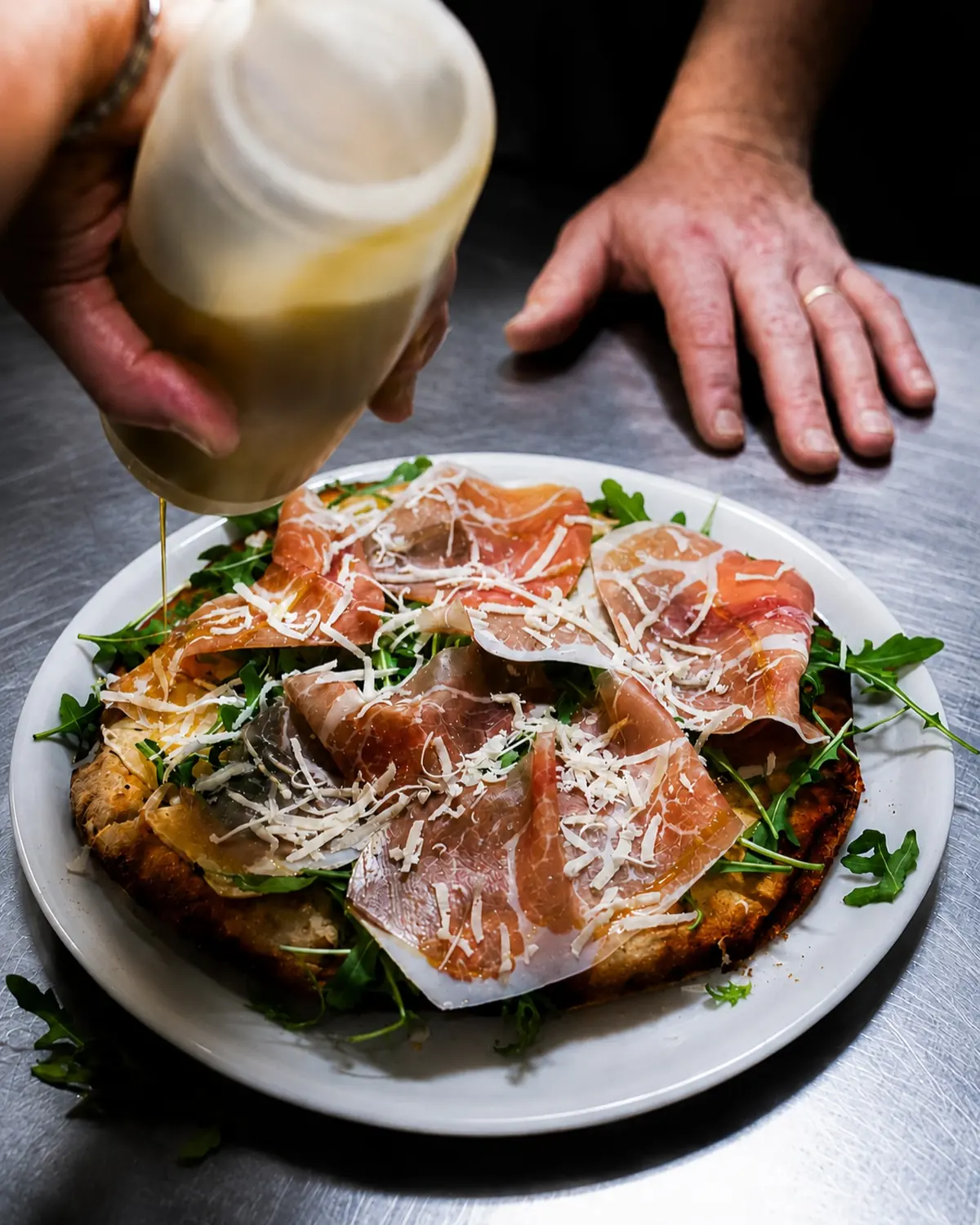 Il pizzaiolo versa un filo d'olio su una pizza con prosciutto crudo, rucola e scaglie di grana