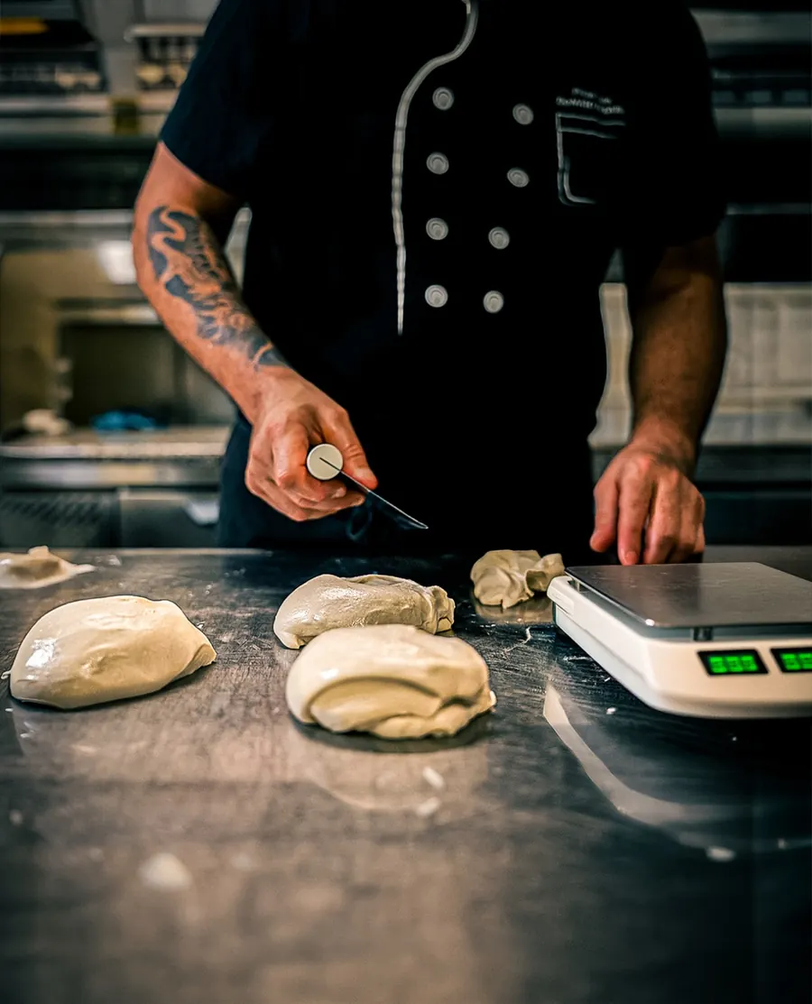 Il pizzaiolo divide l'impasto in panetti sul bancone d'acciaio, accanto alla bilancia